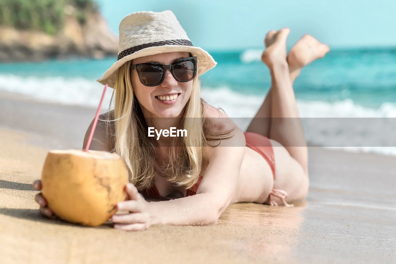 Portrait of smiling young woman in sunglasses,  in summer hat laying on sea beach and drink coconut.