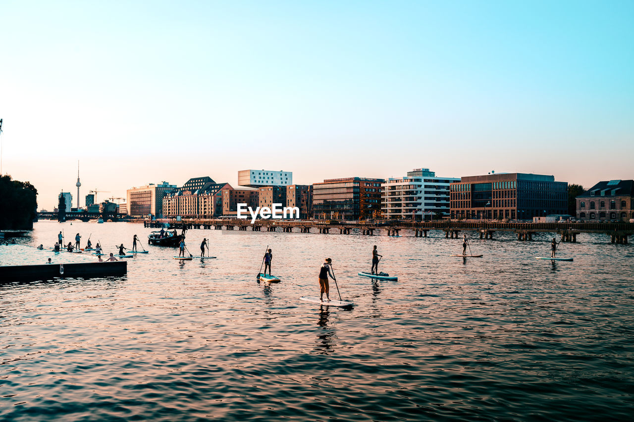 People paddleboarding in river by buildings in city against clear sky during sunset