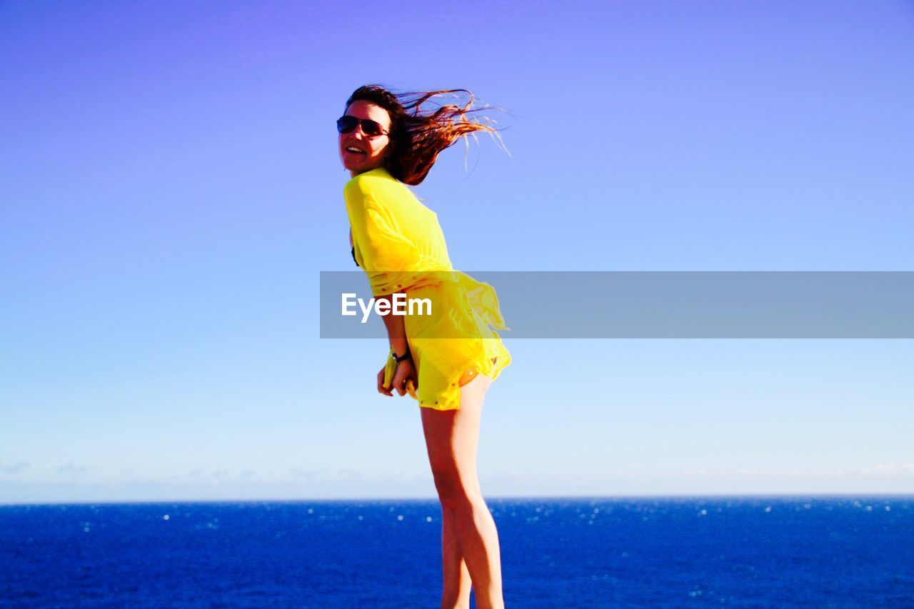 Side view of happy woman in yellow top standing at beach against sea and sky on sunny day