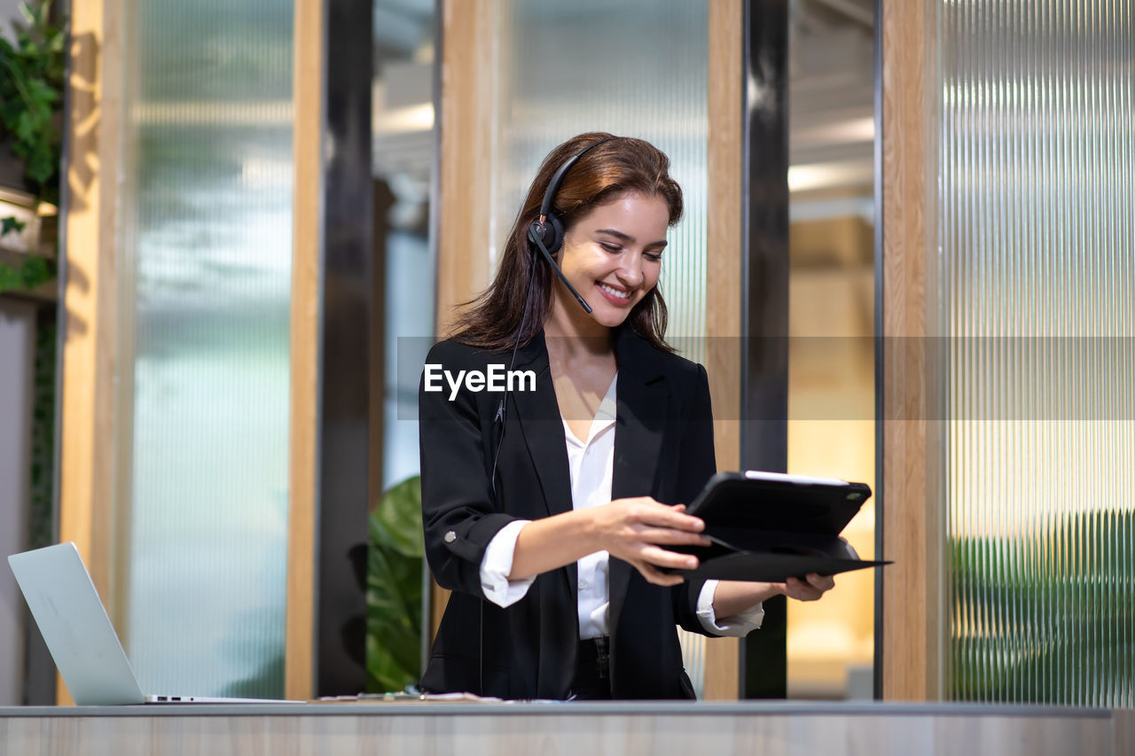 SMILING YOUNG WOMAN USING PHONE WHILE LOOKING THROUGH CAMERA AT HOME