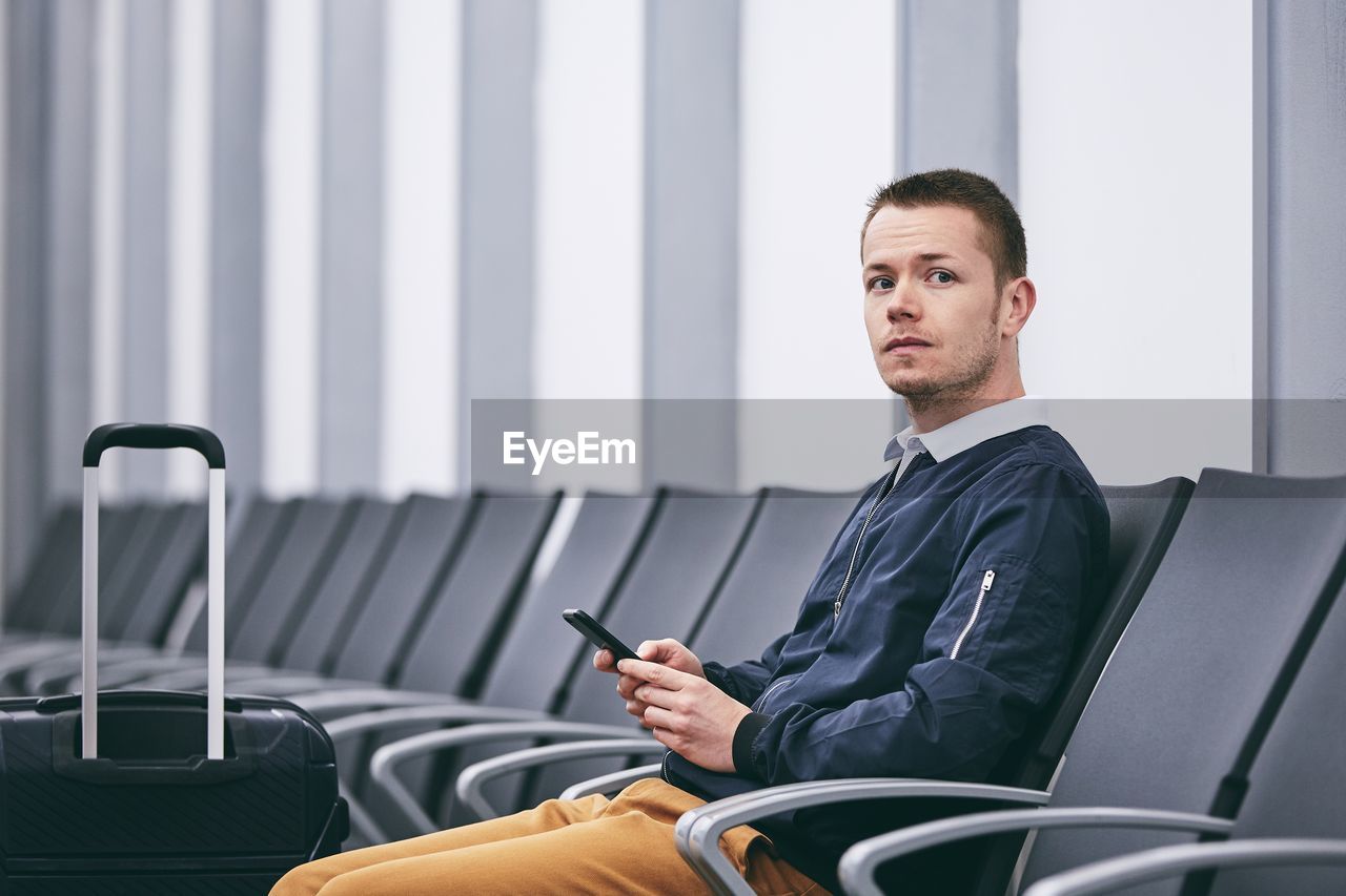 Portrait of man using mobile phone while sitting on chair at airport terminal