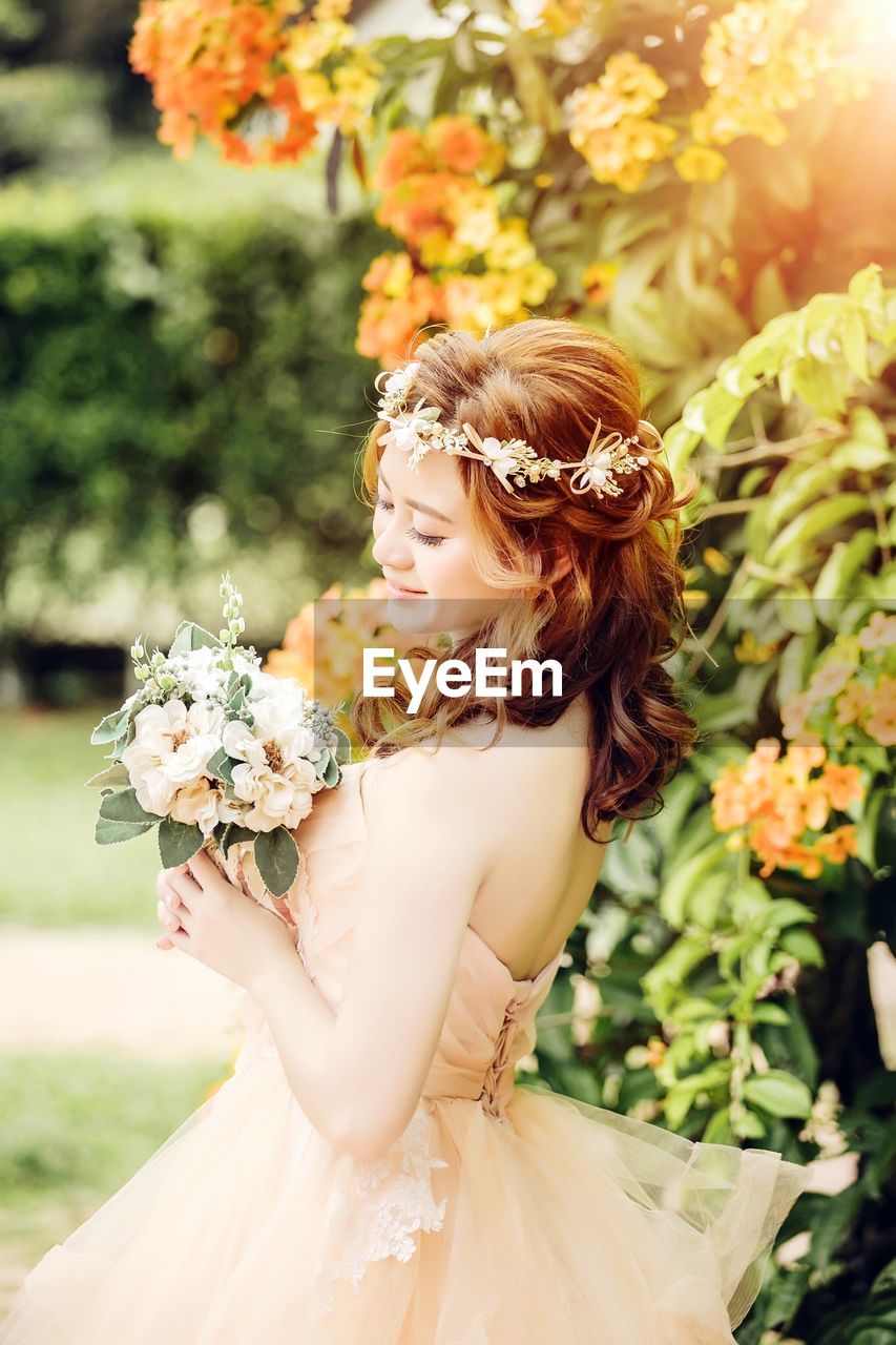 Side view of beautiful bride holding bouquet while standing by plants at park