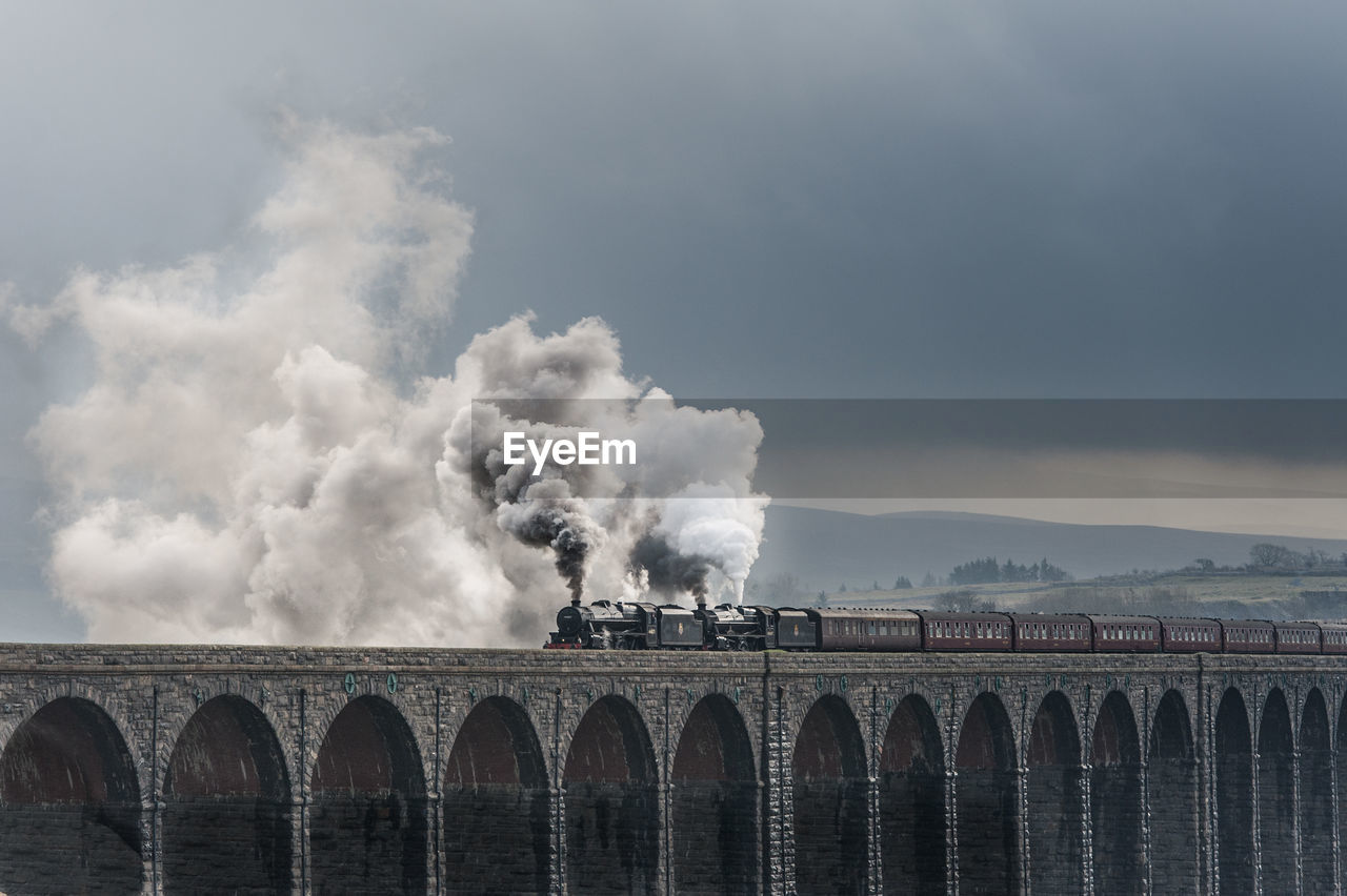 Smoke emitting from steam train on bridge against cloudy sky