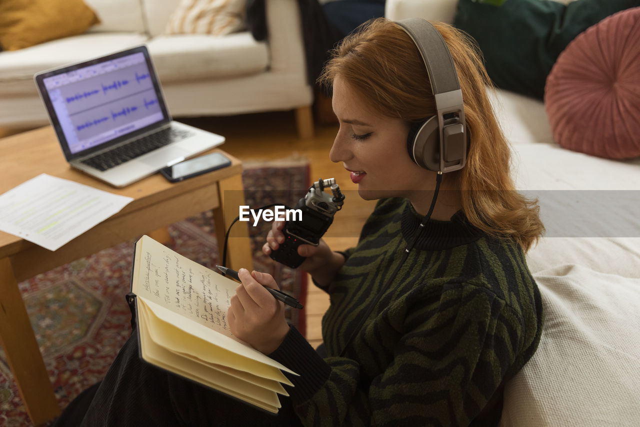 Side view of focused female radio host with mic and headphones writing in notepad while preparing for recording podcast at home