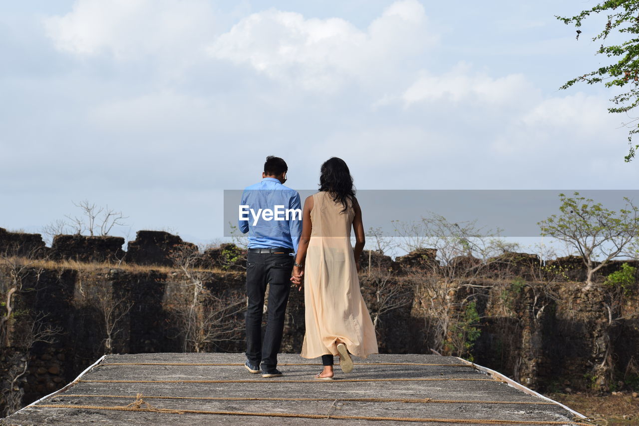 Rear view of couple holding hands while walking on boardwalk against sky