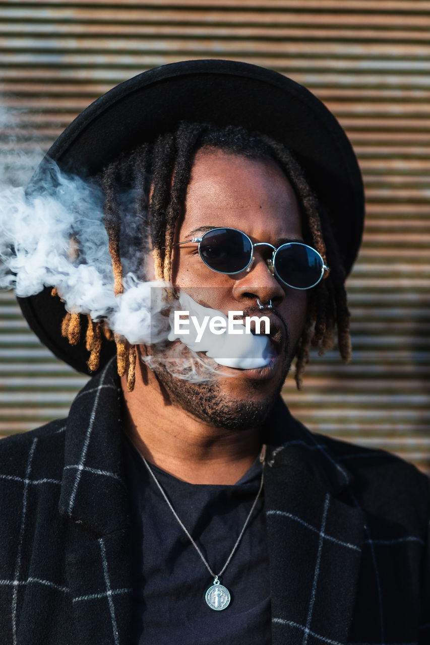 Modern african american man with dreadlocks wearing hat with sunglasses and exhaling smoke on street