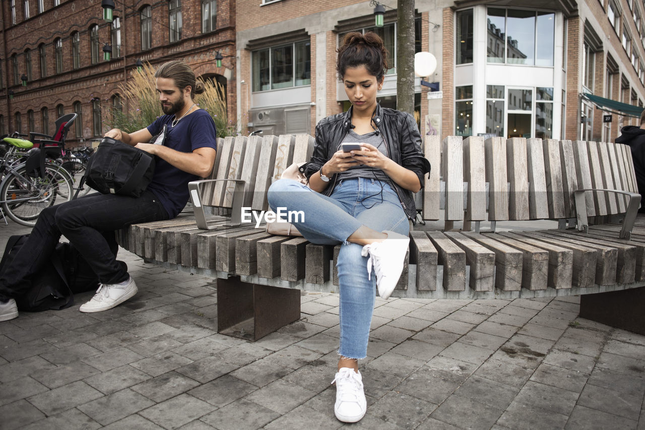 Low angle view of man and woman sitting on wooden bench against building in city