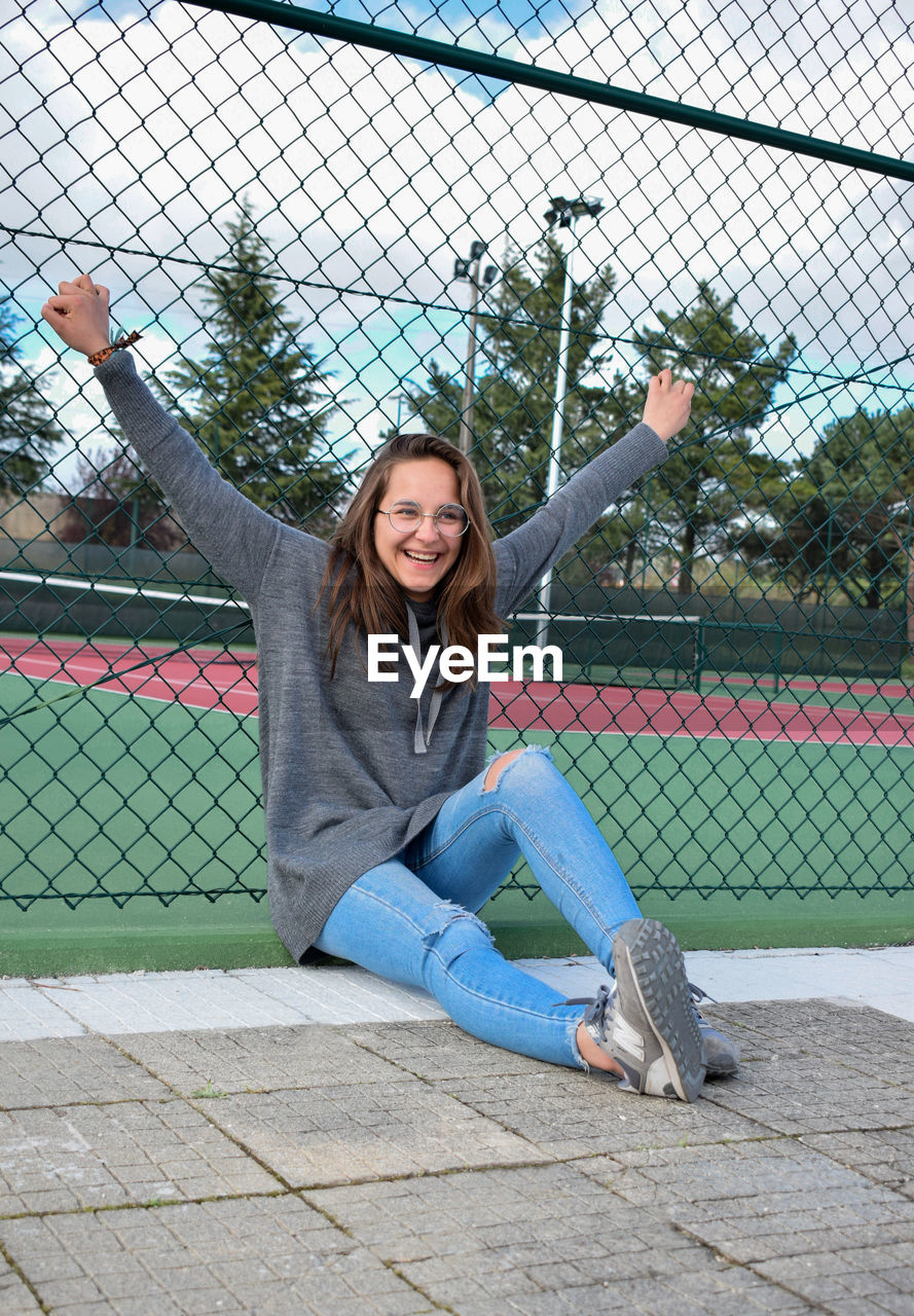 Happy teenage girl sitting against fence at court