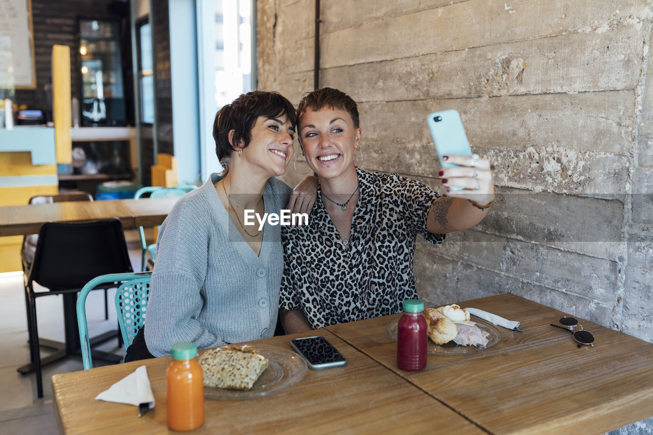 Young friends smiling while taking selfie through mobile phone sitting at restaurant