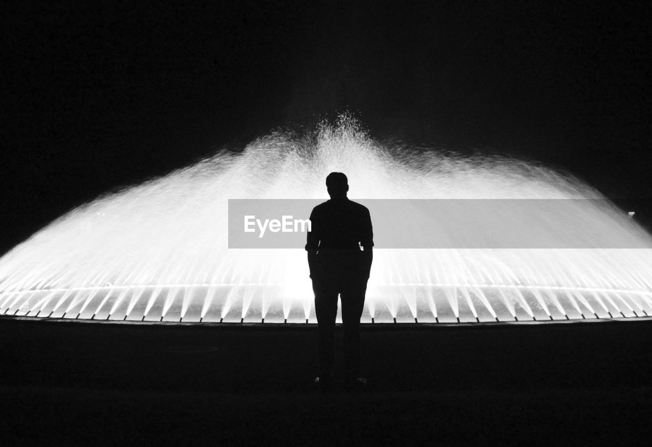Rear view of silhouette man standing in front of fountain at night