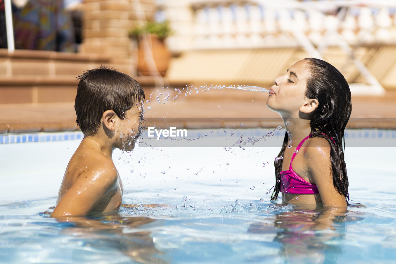 Sister spitting water on brother in swimming pool