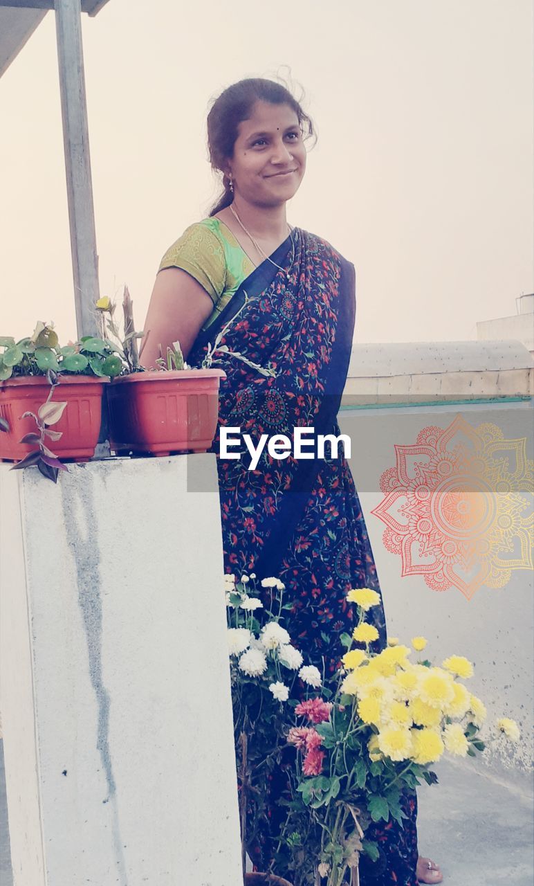 one person, spring, adult, flower, women, nature, dress, standing, flowering plant, blue, portrait, looking at camera, art, plant, clothing, smiling, yellow, young adult, three quarter length, lifestyles, holding, emotion, day, happiness, fashion, person, outdoors, front view, female, architecture, casual clothing, red, leisure activity, multi colored