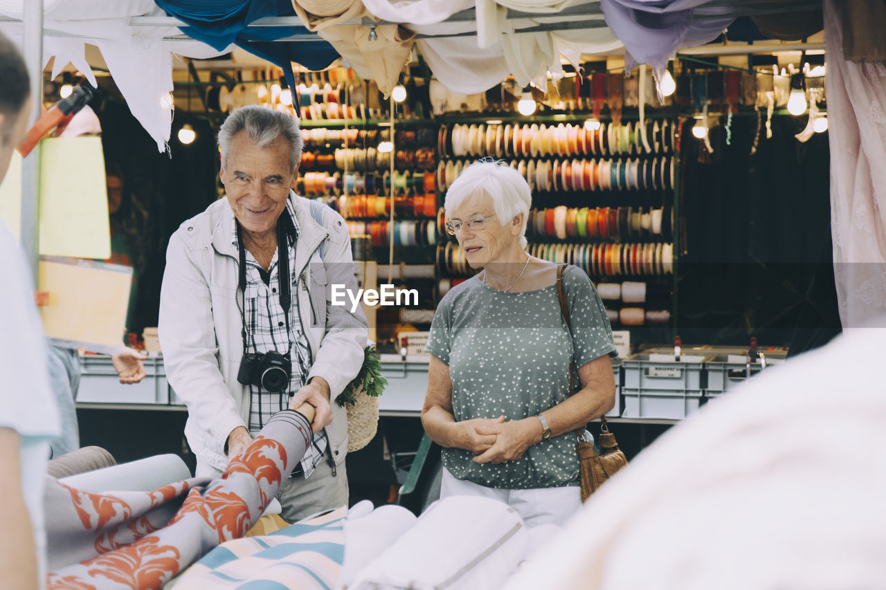 Senior man holding pattern fabric while shopping with woman at market in city