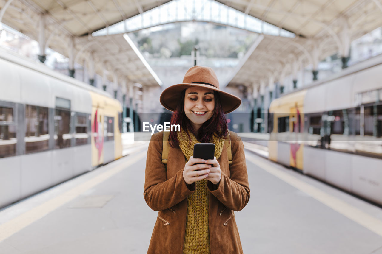 Smiling woman using smart phone while standing at railroad station platform