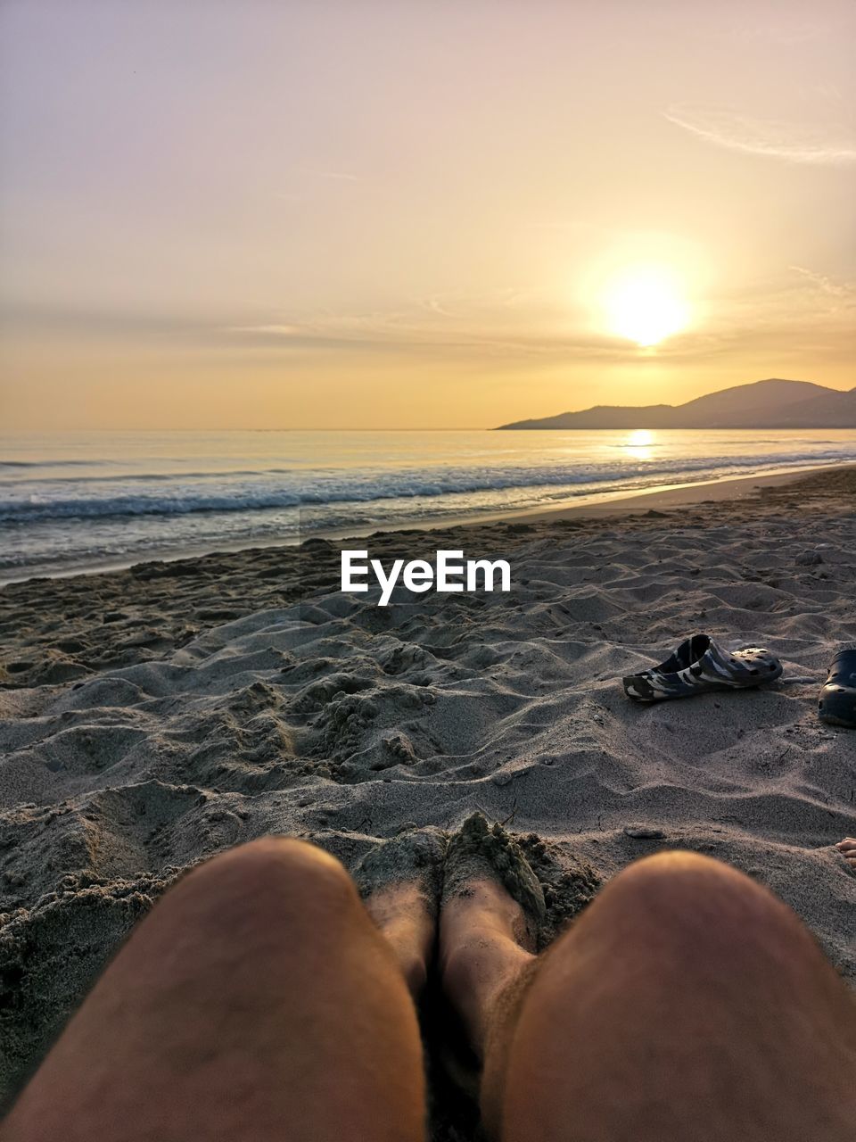 LOW SECTION OF MAN RELAXING ON BEACH AGAINST SKY DURING SUNSET