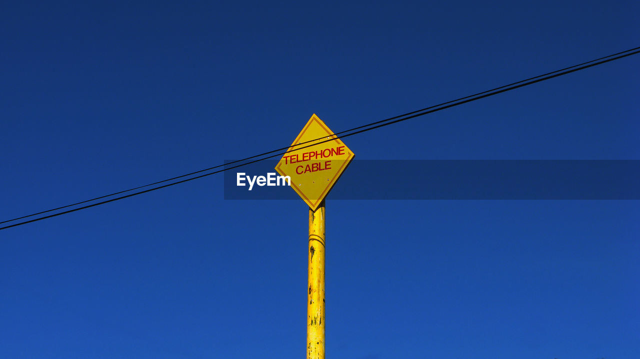 Low angle view of sign board by cable against clear blue sky