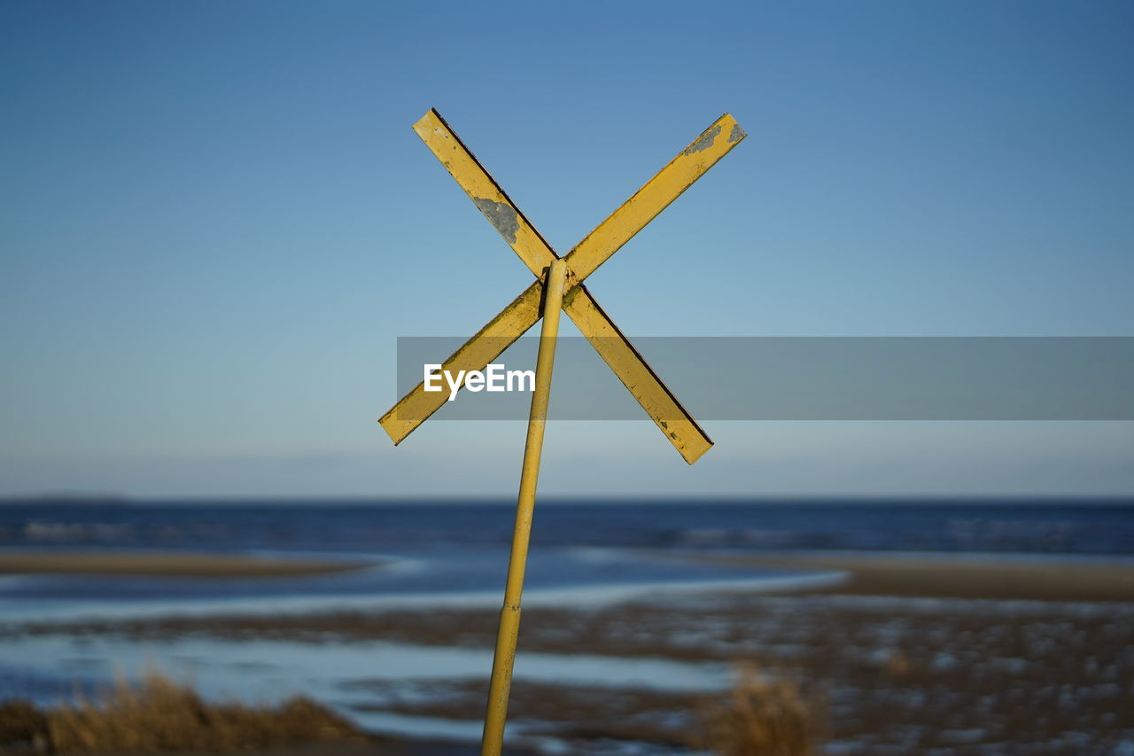 Yellow warning cross at coast for nautical navigation on beach against clear sky