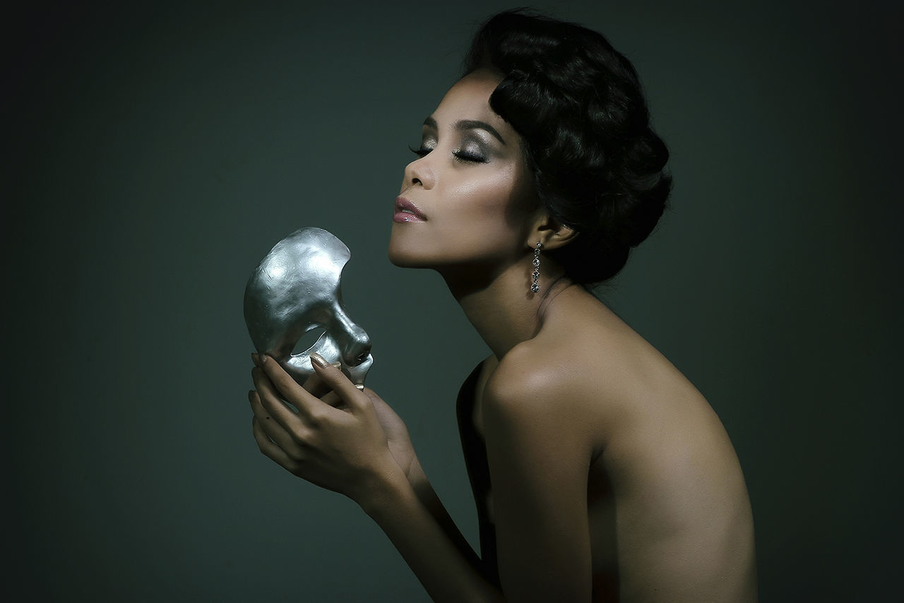 Side view of naked fashion model holding silver mask against gray background