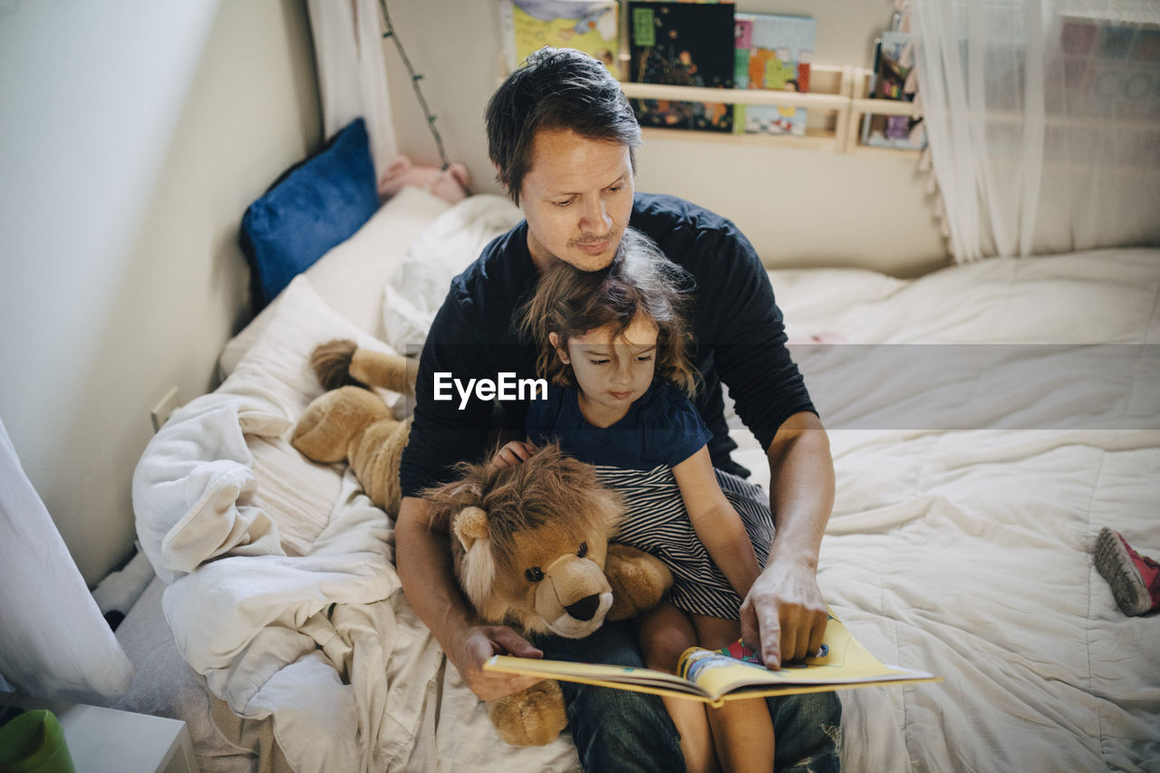 High angle view of father reading book with daughter while sitting in bedroom