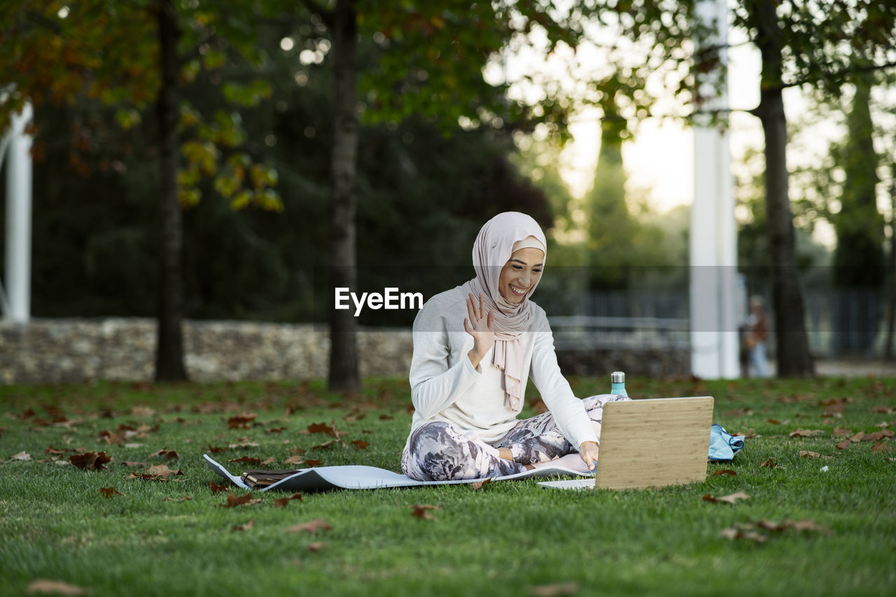 Full body of cheerful arab female in hijab waving hand while having video chat via laptop on grassy lawn in park
