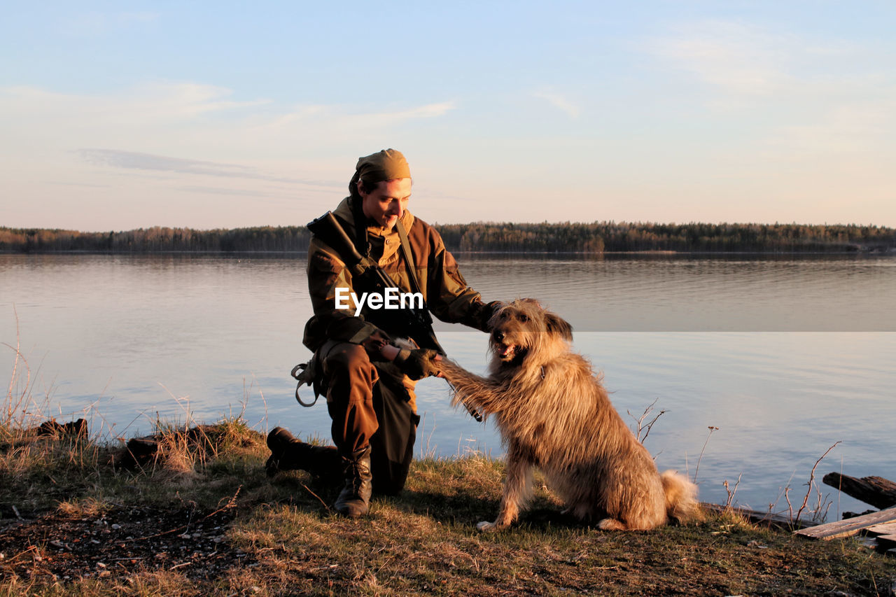 A man in a camouflage suit sitting on the shore of a lake with a dog