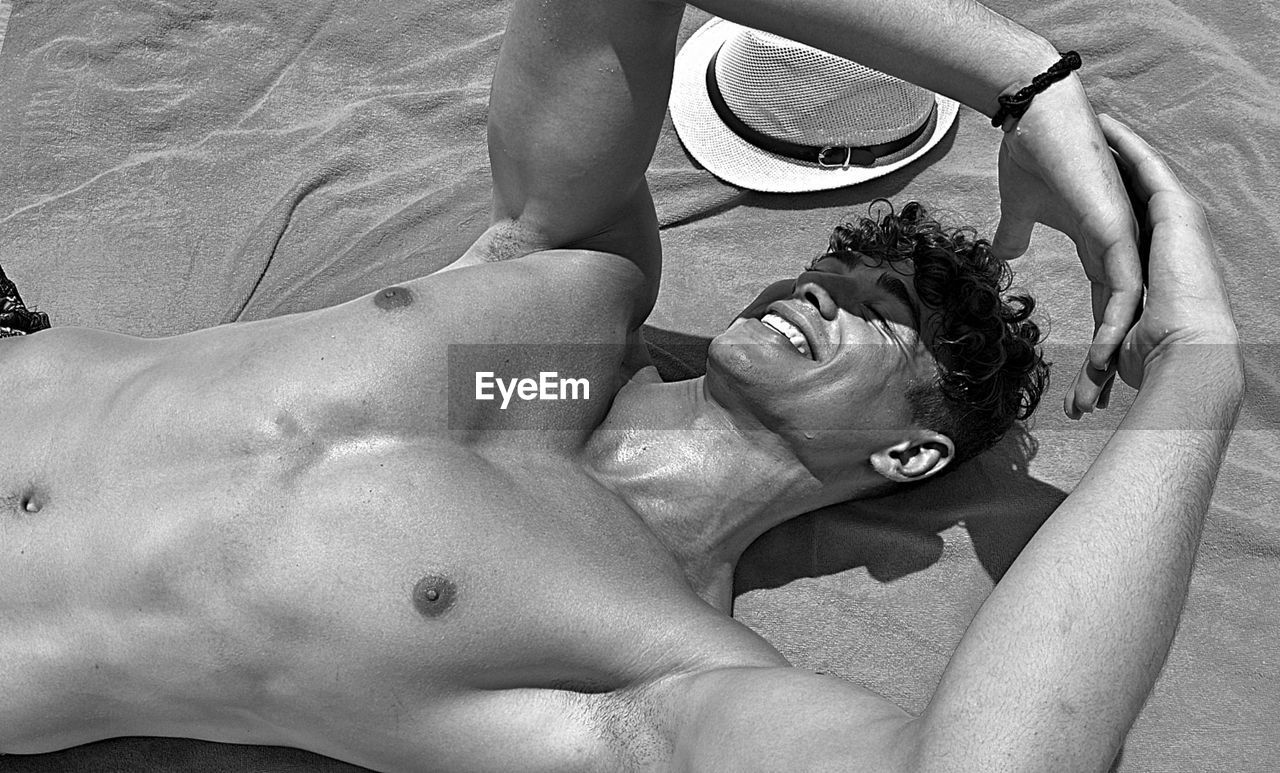 Shirtless man smiling while relaxing at beach in sunny day