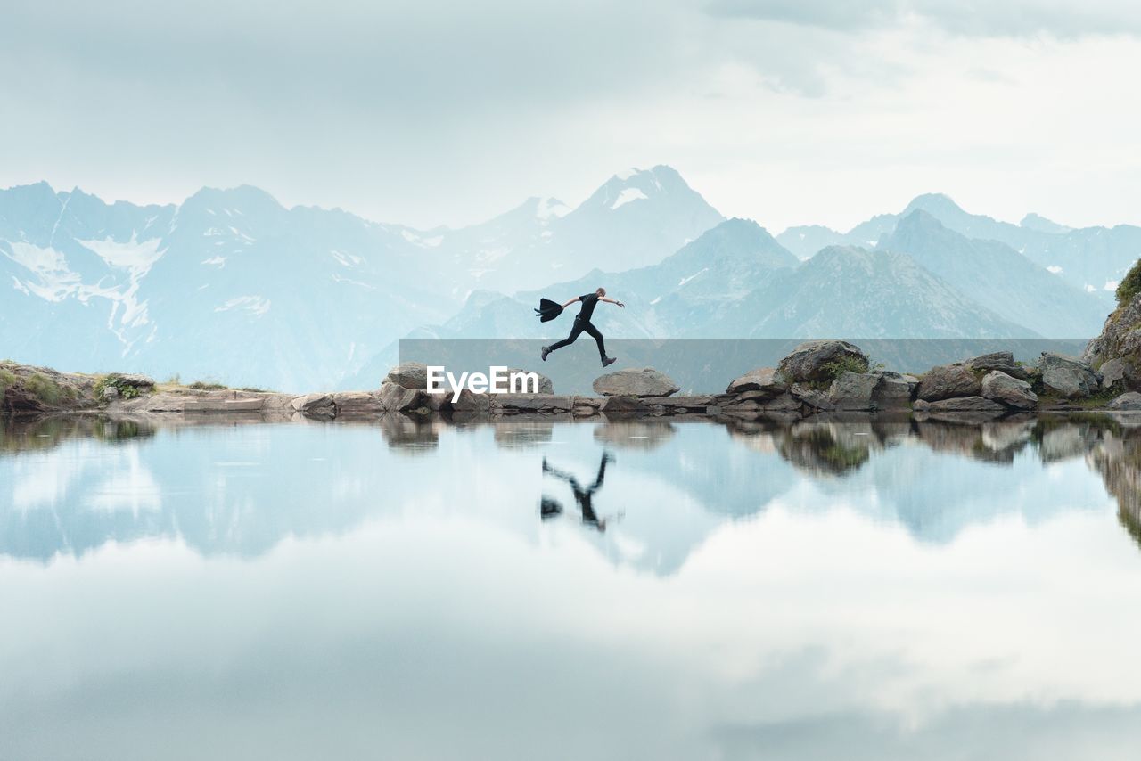 Man reflecting on calm lake while jumping mountains against cloudy sky