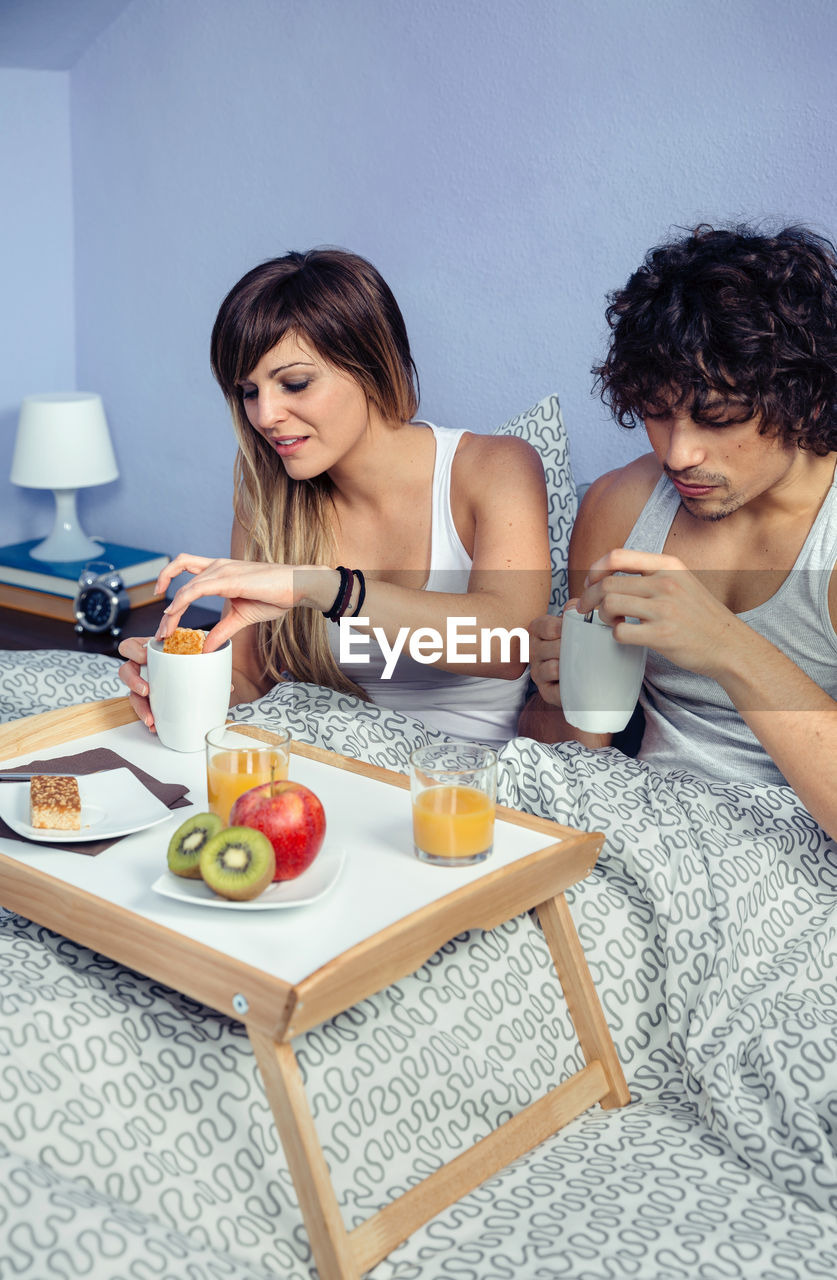 Young couple having food and drinks on bed at home