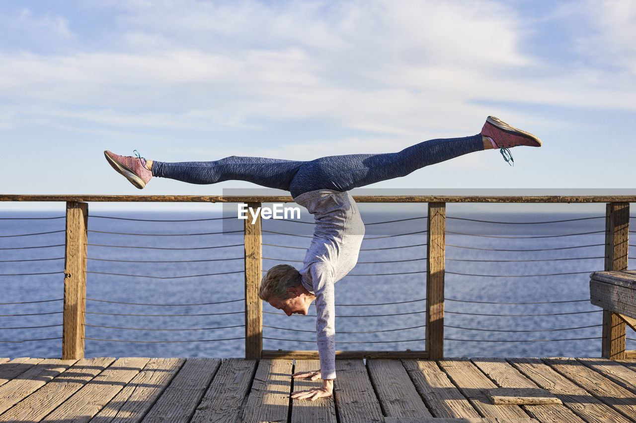 Full length senior woman doing handstand while exercising on floorboard by sea against sky