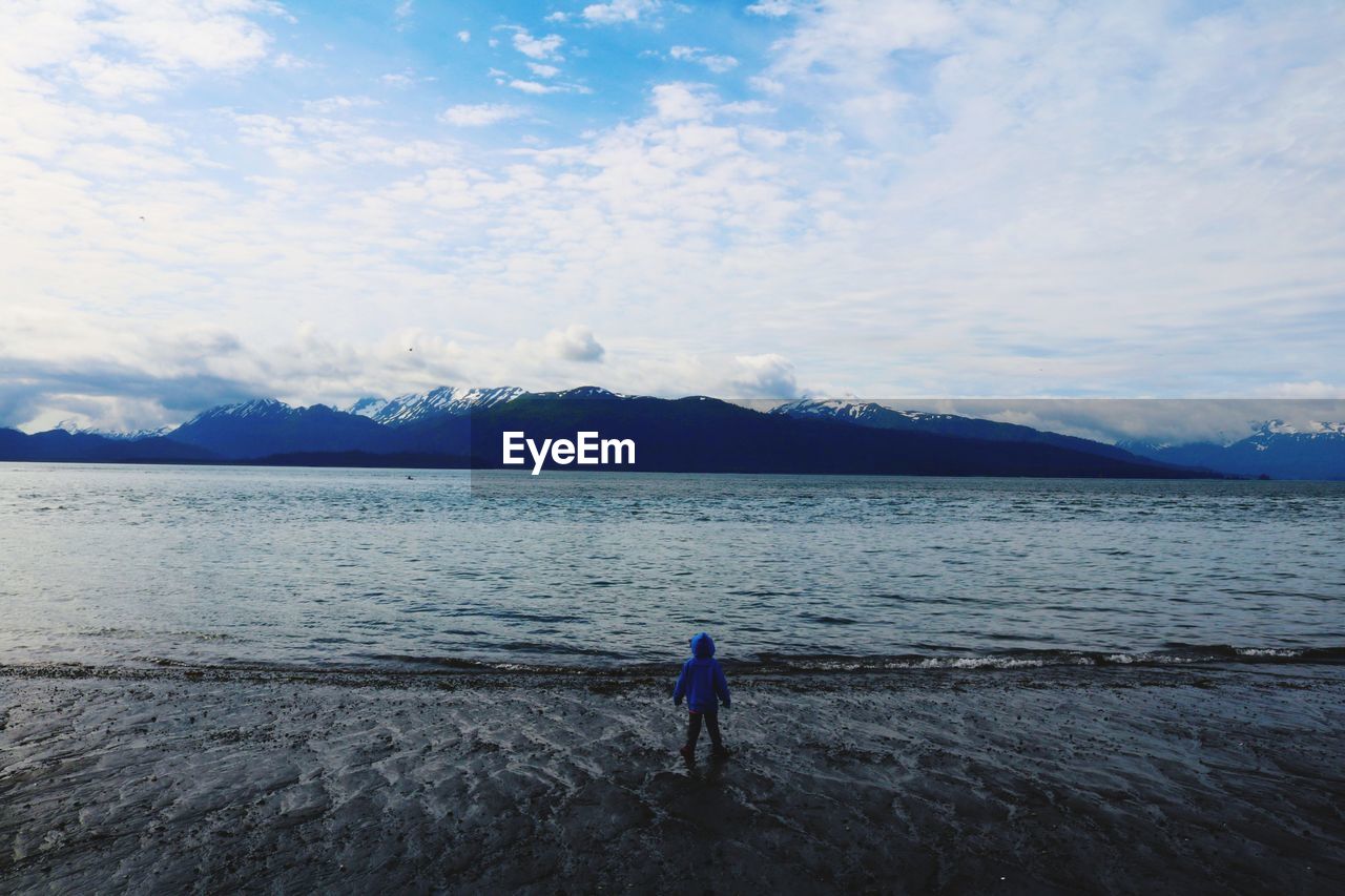 Rear view of child standing on lakeshore against sky