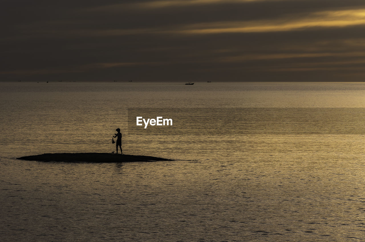 Silhouette man standing in sea against sky during sunset