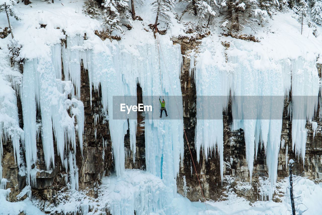 Panoramic view of snow-covered land and trees in forest with ice climber on frozen waterfall.