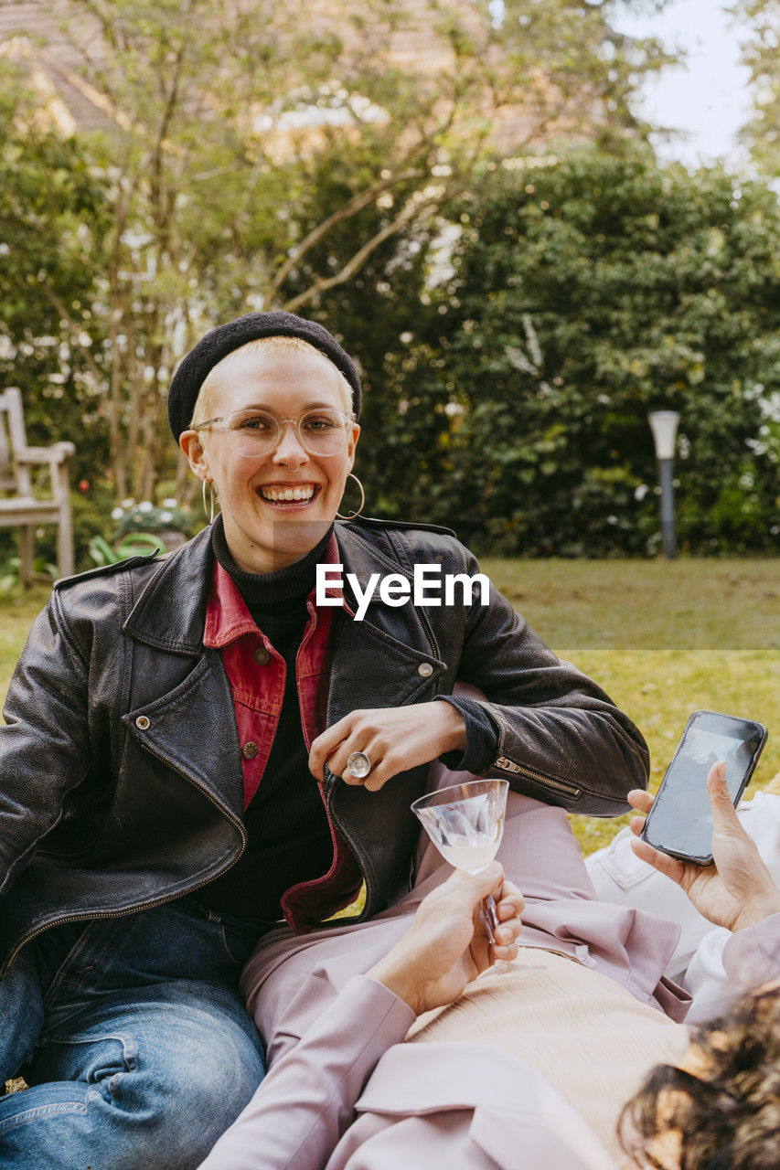 Portrait of happy gay woman wearing leather jacket while sitting with friend in back yard