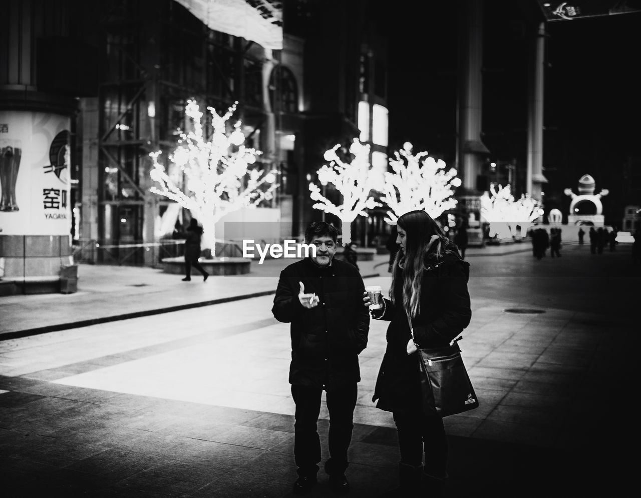 PEOPLE WALKING ON ILLUMINATED STREET IN CITY AT NIGHT