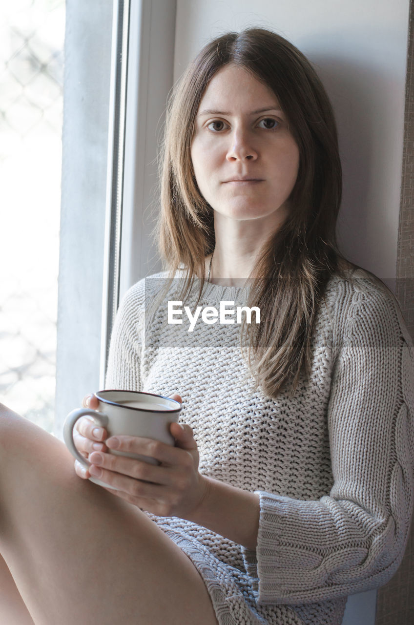 Beautiful young woman sitting near the window with a cup of coffee in her hands