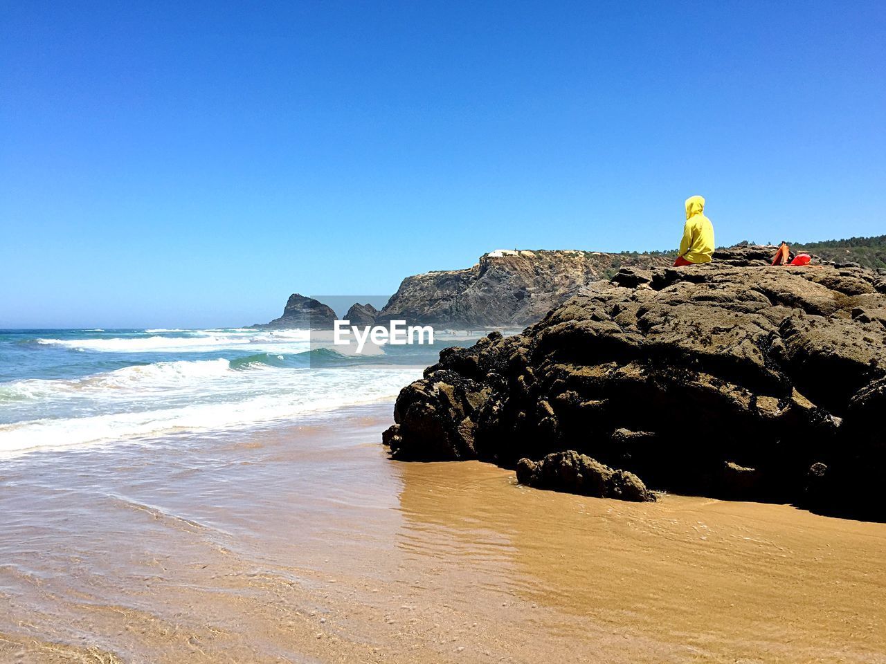 Person sitting on rock at beach against clear blue sky