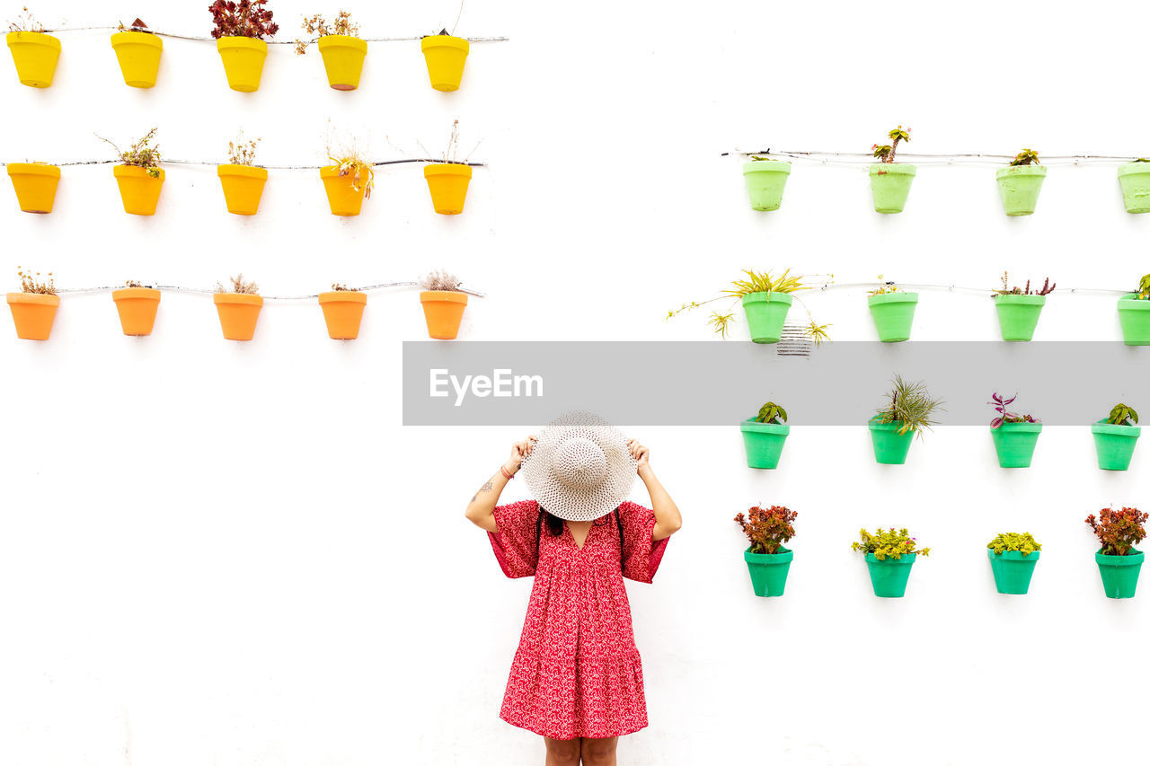 Anonymous female tourist covering face with straw hat near white wall with different potted plants