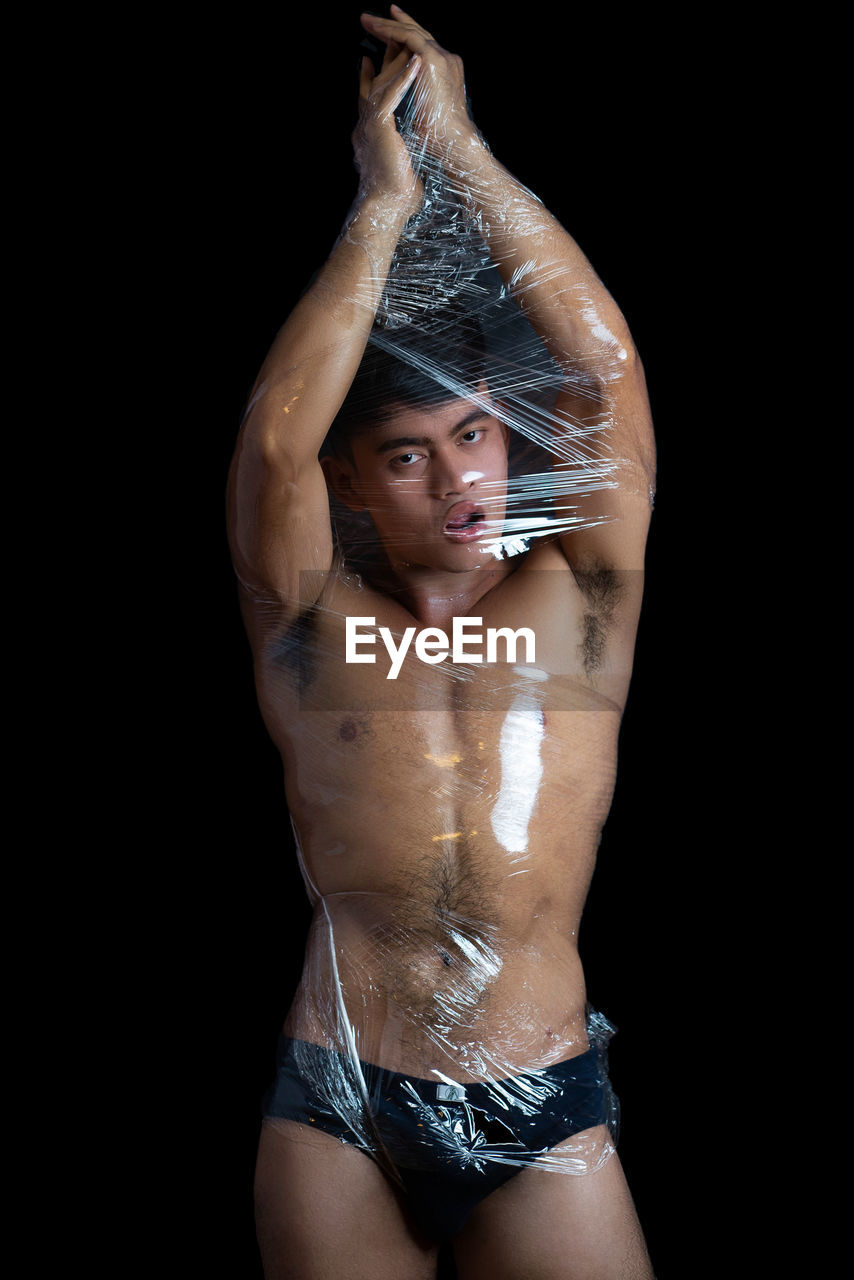 Portrait of shirtless young man wrapped in plastic against black background