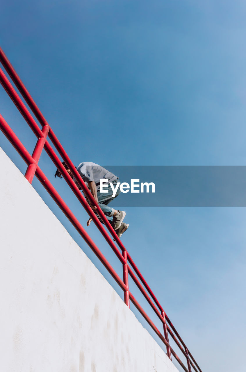 From below of brave male jumping over metal fence in street and showing parkour trick against blue sky
