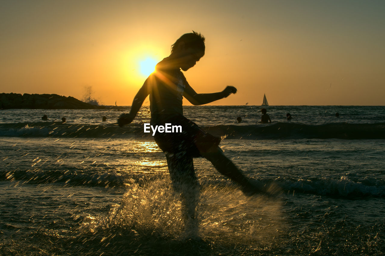 Boy kicking water in sea during sunset