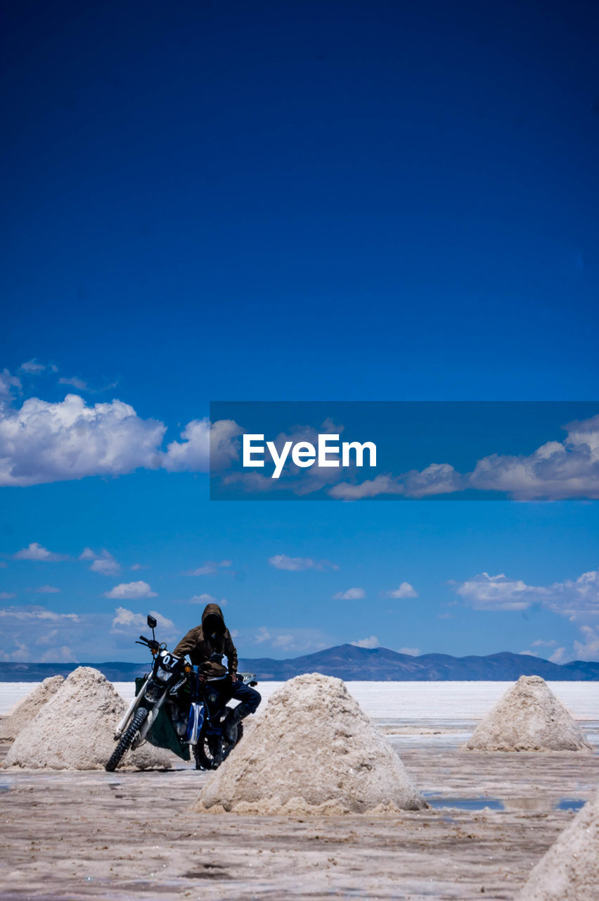 Hooded person with motorcycle on salt flat at salar de uyuni against sky