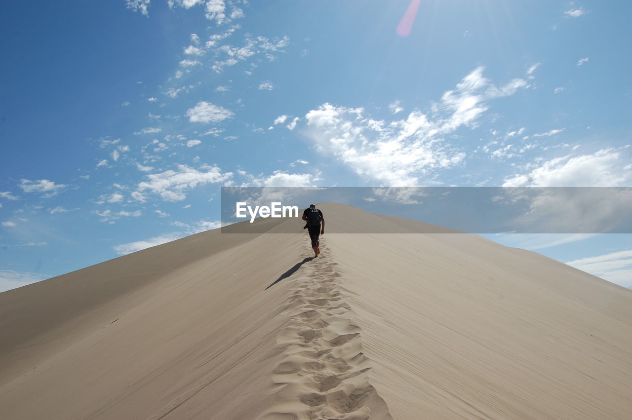 Rear view of man climbing on sand dune at desert against sky during sunny day