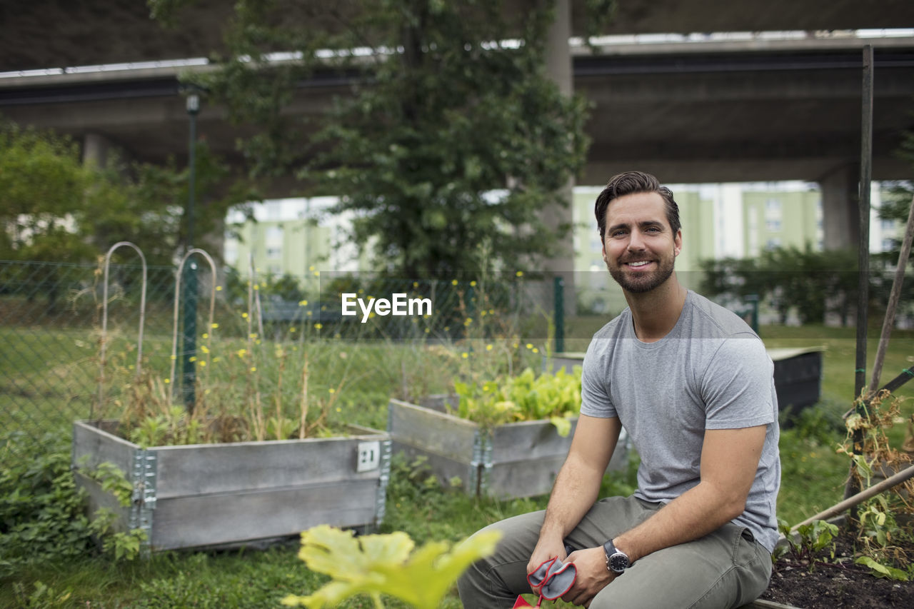 Portrait of smiling mid adult man sitting on edge of wooden crate at urban garden