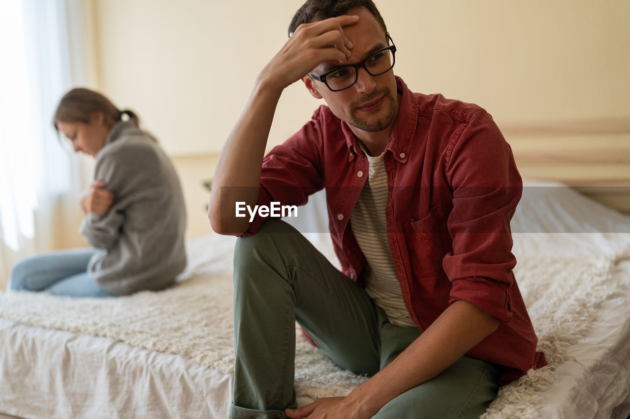 Young unhappy family couple sitting separately on bed at home, having problems in relationship