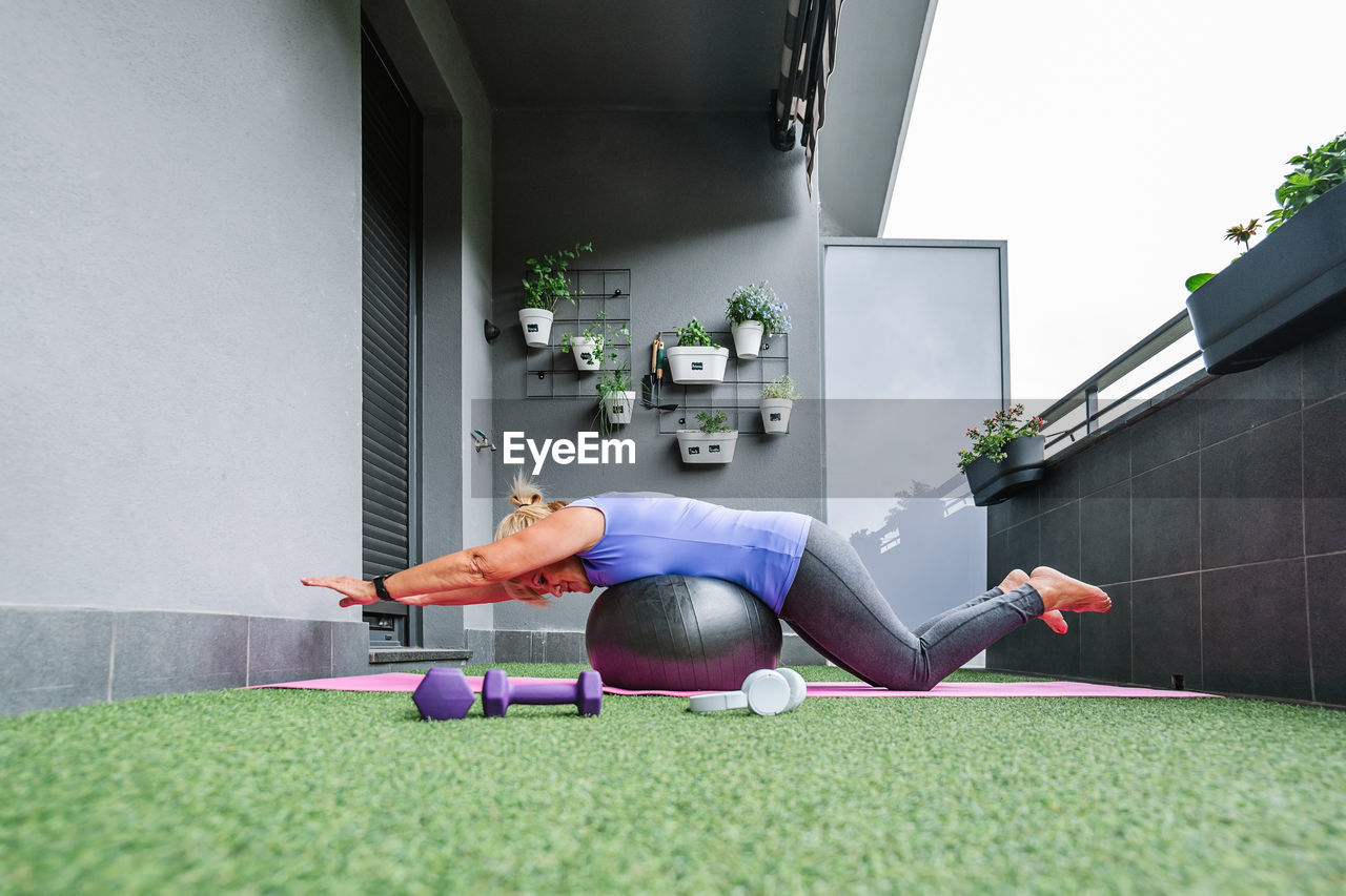 Full length profile of elderly woman in sportswear balancing on fit ball at balcony