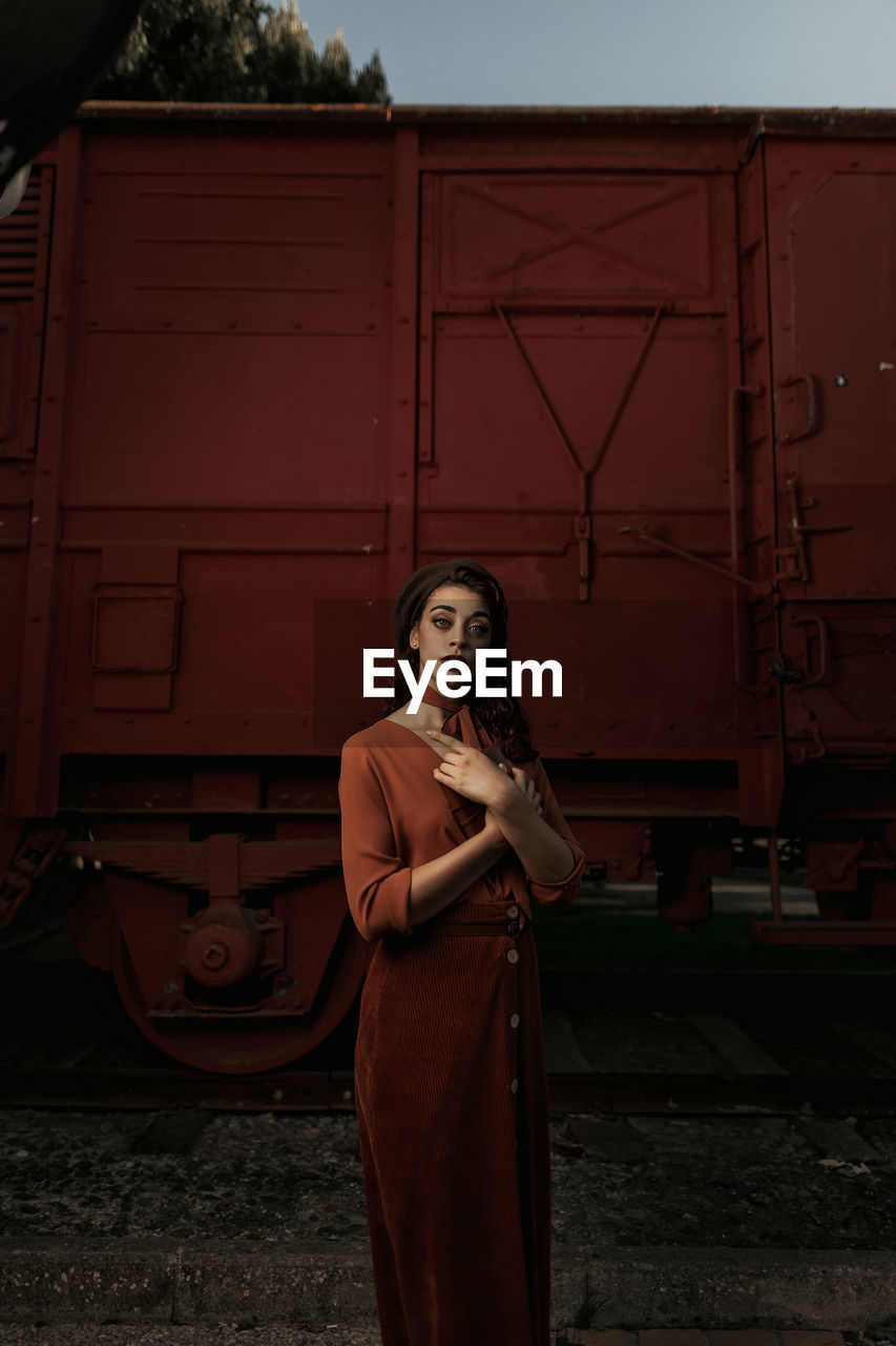 Dark haired female totally wearing terracotta clothes in vintage style standing near terracotta car train and holding open book in hands having dreaming look