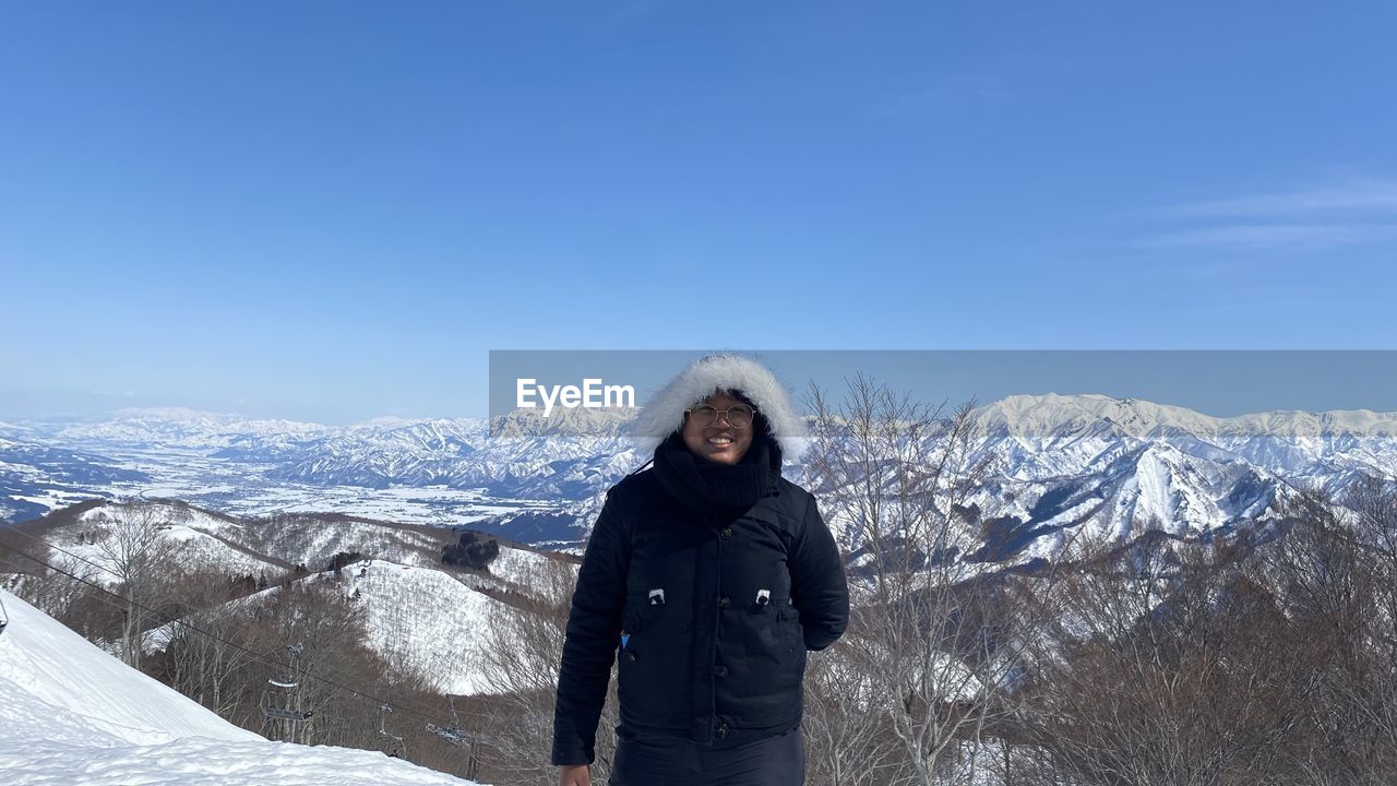 portrait of young woman standing on snowcapped mountain against clear blue sky