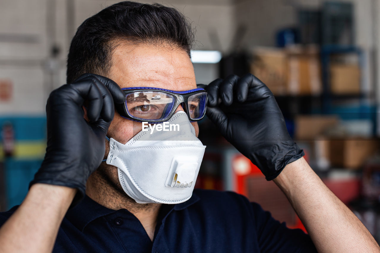 Adult man in latex gloves and respirator putting on protective goggles and looking at camera during work in garage