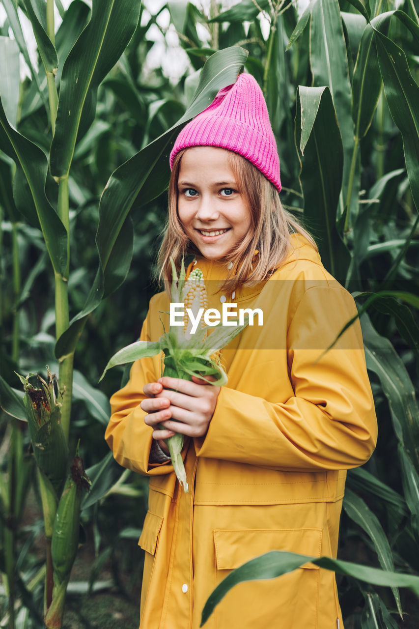 Stylish teenage girl in yellow raincoat and hot pink hat laughing on corn field