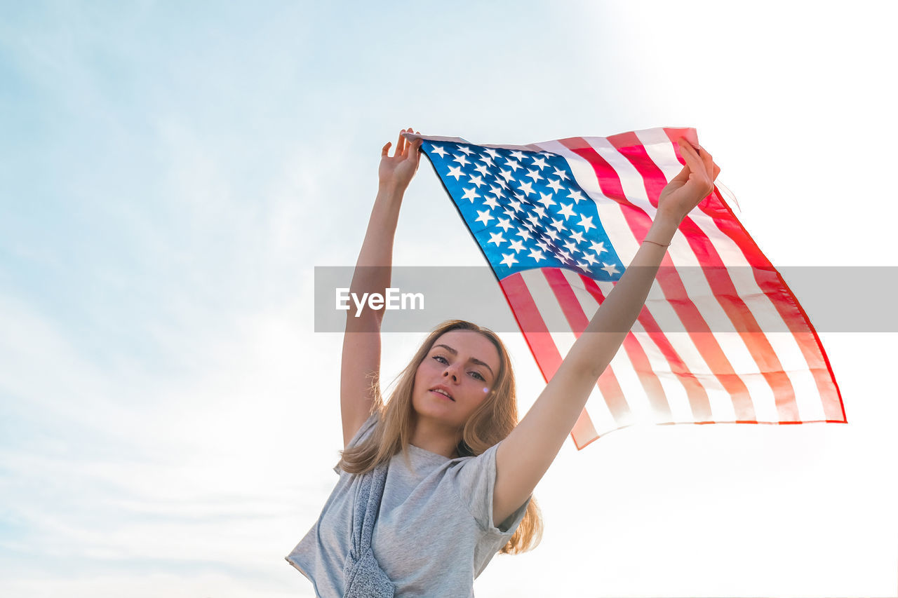 Young millennial blonde woman standing with american flag next to blue sky. flag of the united 